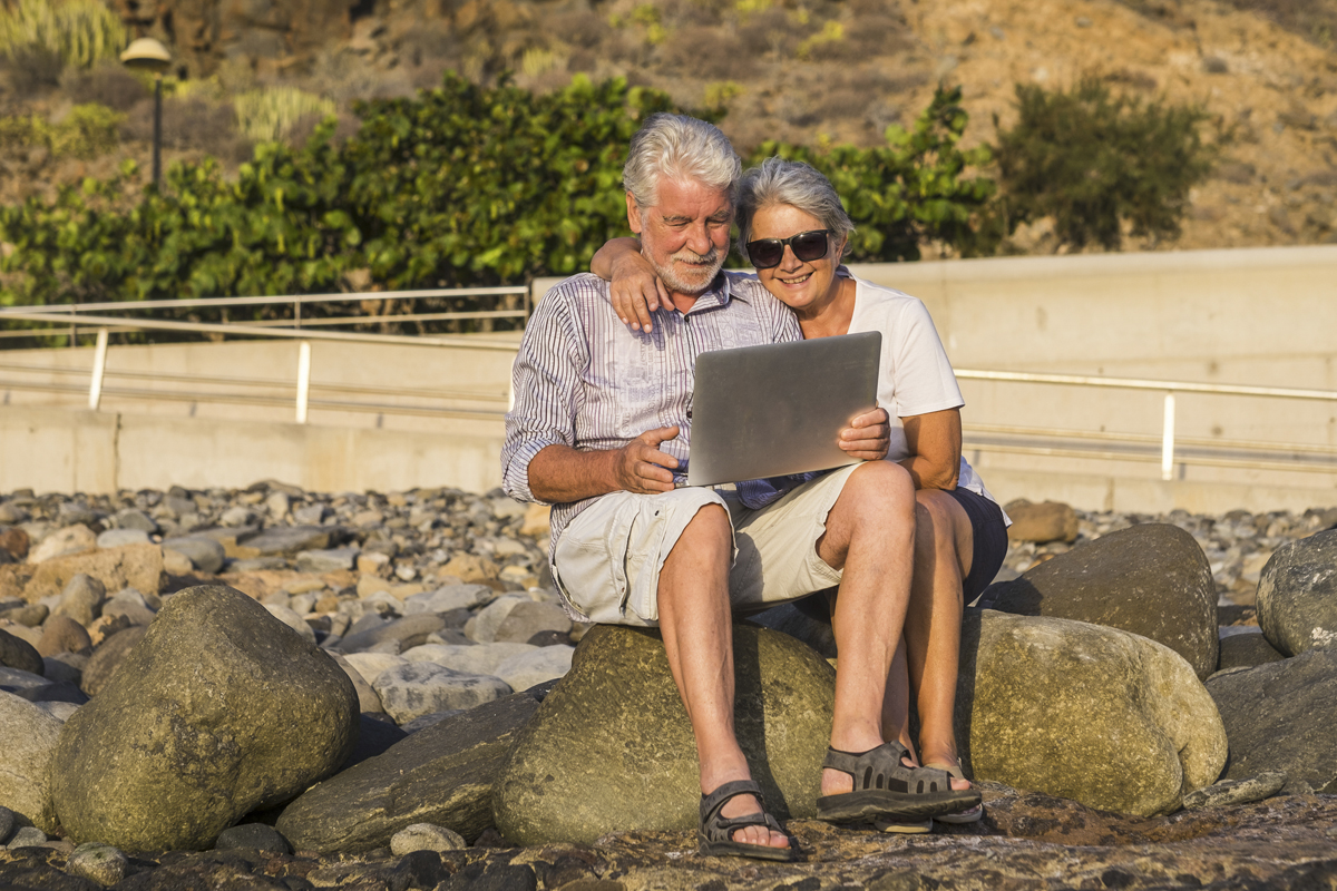 concept of vacation, technology, tourism, travel and people – happy senior couple with tablet pc computer on pebble beach. White hair