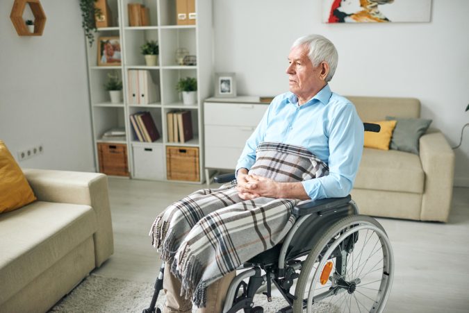 Sad and serene senior disable man sitting in wheelchair by couch at home