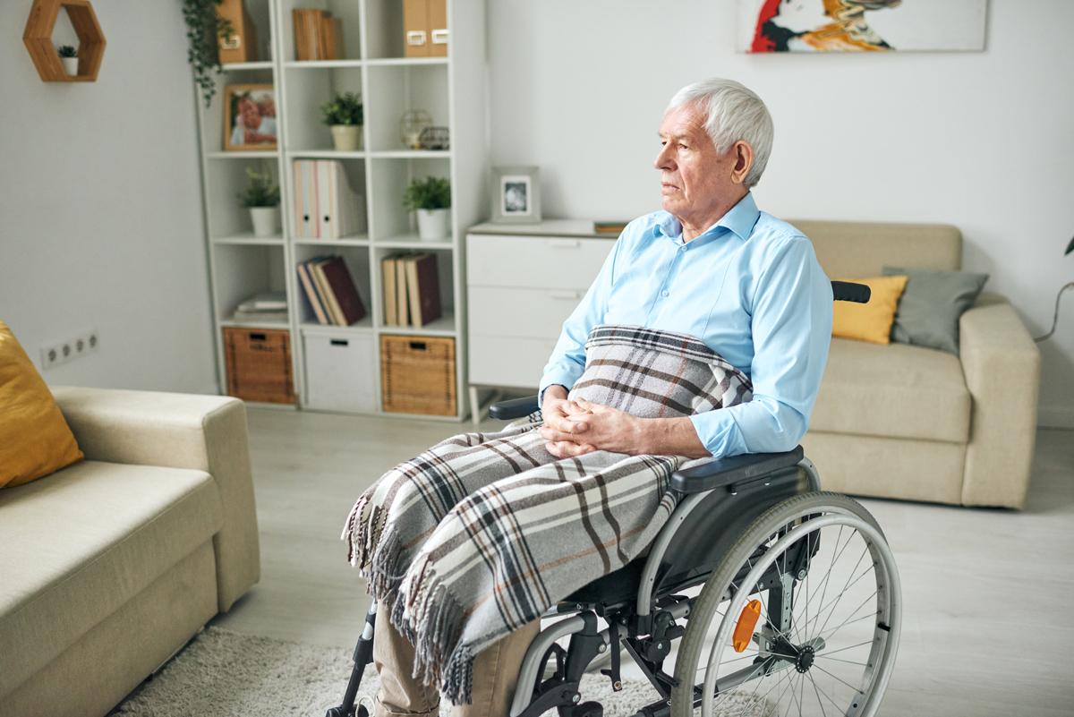 Sad and serene senior disable man sitting in wheelchair by couch at home
